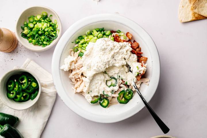 Bowl with shredded chicken, diced tomatoes, spring onions, jalapeño slices, topped with creamy ricotta. Nearby bowls hold more ingredients.